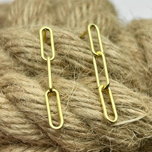 May include: Gold-colored chain link earrings. Each earring features a series of elongated oval links connected to form a dangling design. The earrings are displayed on a textured, neutral-toned surface.