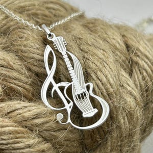 Op de afbeelding: Zilverkleurige hangerketting met een gitaar en een G-sleutel ontwerp. De gitaar is gedetailleerd met snaren en een klankgat. De hanger hangt aan een fijne ketting, voor muziekliefhebbers.