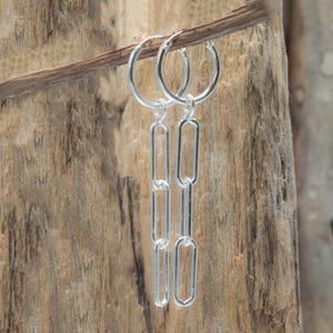 May include: A pair of silver hoop earrings with dangling rectangular chain links. The earrings are displayed against a natural wood background, highlighting their bright, reflective surface. The design is simple and elegant.
