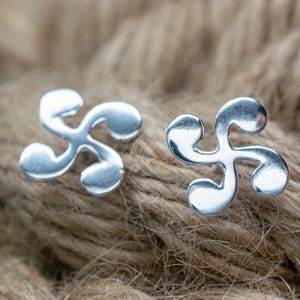 May include: A pair of silver-colored stud earrings. Each earring features a stylized, three-lobed design. The earrings are displayed on a textured, brown surface, creating a contrast with their shiny appearance. The design is reminiscent of a Basque Lauburu.