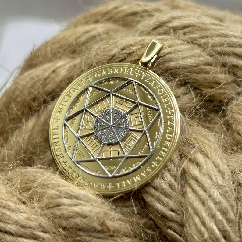 May include: Gold-colored pendant with intricate geometric designs and text. The pendant features a central silver-colored circle surrounded by a star-like pattern. The outer ring has engraved text. The pendant is suspended from a gold-colored bail.