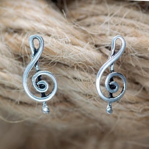 Könnte beinhalten: Ein Paar silberfarbene Ohrringe in Form eines Notenschlüssels. Jeder Ohrring zeigt das ikonische Musiksymbol mit abgerundeter Basis und geschwungenem Oberteil. Die Ohrringe werden vor einem strukturierten, neutral getönten Hintergrund präsentiert.