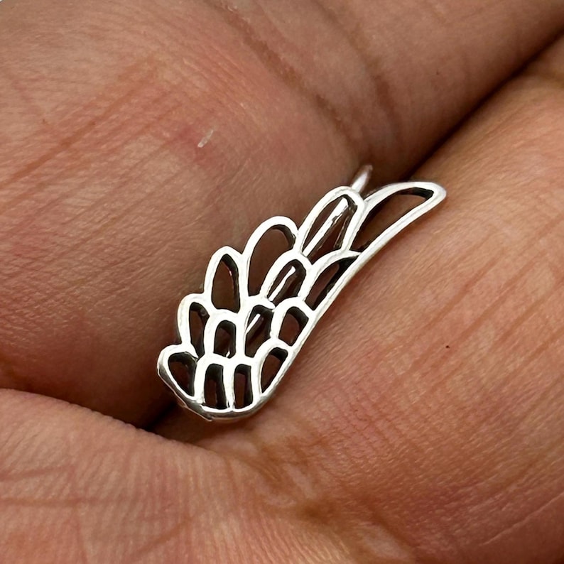 May include: A small, silver-colored wing-shaped pendant. The pendant has a detailed design with open spaces, giving it a delicate appearance. The wing is slightly curved and has a loop at the top for attaching to a chain or other jewelry.