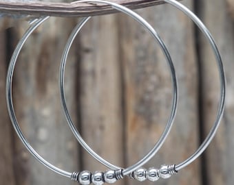 May include: A pair of silver hoop earrings. Each earring features a series of small, round beads clustered near the bottom. The earrings are displayed against a blurred wooden background.