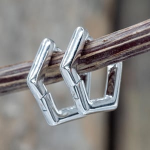 May include: Two silver square-shaped earrings with a hinged design are displayed. The earrings are hanging on a thin, brown twig. The background is blurred, suggesting an outdoor setting. The earrings have a polished, reflective surface.