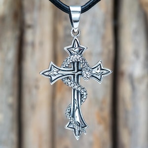 Könnte beinhalten: Silberfarbener Anhänger mit einem Kreuz-Design, das von einer Schlange umwunden ist. Das Kreuz hat spitze Enden und die Schlange ist detailliert. Der Anhänger hängt an einem schwarzen Lederband, was einen gotischen Look erzeugt.