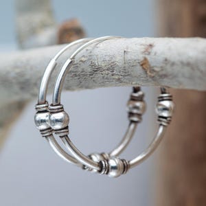 May include: Silver hoop earrings with multiple round beads. The earrings are hanging from a branch.