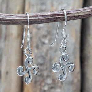 May include: A pair of silver-colored dangle earrings. Each earring features a Celtic triskele design, suspended from a simple hook. The triskeles are three-pronged, with a swirling, curved shape. The earrings are displayed against a natural, wooden background.