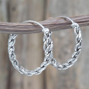 Puede incluir: Un par de pendientes de aro plateados con un diseño trenzado. Los pendientes son redondos y tienen un aspecto texturizado y tejido. Los pendientes se muestran sobre un fondo de madera borroso.