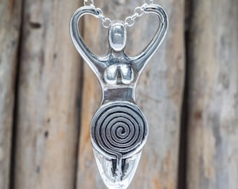 May include: A silver pendant necklace featuring a stylized female figure with arms raised. The pendant has a spiral design on the lower portion and is suspended from a delicate chain. The background is a blurred wooden surface.