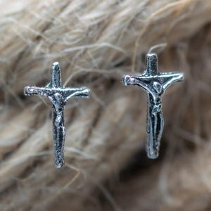 Könnte beinhalten: Zwei silberfarbene Kreuz-Ohrstecker. Jeder Ohrring zeigt eine detaillierte Darstellung von Jesus am Kreuz. Die Ohrringe sind vor einem strukturierten, neutral getönten Hintergrund platziert.