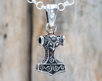 Op de afbeelding: Een zilveren hanger in de vorm van Thors hamer, Mjolnir, hangt aan een zilveren ketting. De hamer heeft ingewikkelde, donkergekleurde details. De achtergrond is een vage houten ondergrond. Een sieraad.