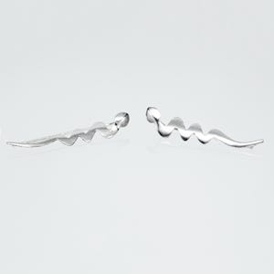 May include: A pair of silver-toned earrings. Each earring features a wavy, elongated design with a small, rounded end. The earrings are displayed against a white background, highlighting their minimalist and modern aesthetic.