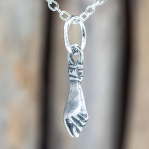 Könnte beinhalten: Eine silberfarbene Anhängerkette mit einem detaillierten Handgesten-Anhänger. Der Anhänger, eine geballte Faust, hängt an einer filigranen Kette. Der Hintergrund ist eine verschwommene Holzoberfläche.