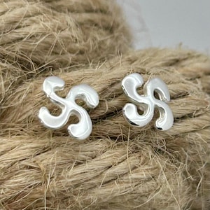 May include: A pair of silver stud earrings featuring a curved, four-armed spiral design known as a lauburu. The earrings have a smooth, polished metallic finish and a symmetrical shape with rounded ends on each of the four curved arms.