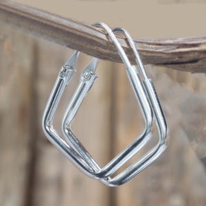 May include: A pair of silver square hoop earrings. The earrings are made of shiny metal and have a simple, geometric design. They are displayed against a blurred wooden background.