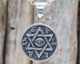 Puede incluir: Un collar con colgante de plata con un dije circular con un diseño de Estrella de David, un ojo y varios símbolos. El colgante cuelga de una delicada cadena. El fondo es una superficie de madera borrosa.