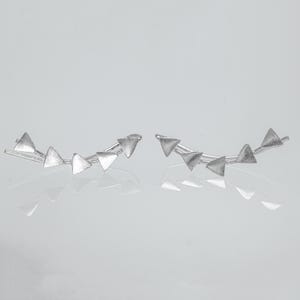 Può includere: Un paio di orecchini a rampicante argentati. Ogni orecchino presenta una serie di piccole forme triangolari disposte lungo una barra curva. Gli orecchini sono esposti su una superficie riflettente, evidenziando il loro design geometrico.