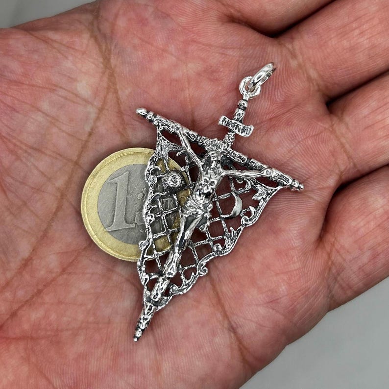 May include: A detailed silver pendant featuring a crucifix design with a filigree pattern. The pendant showcases the figure of Jesus Christ on the cross. A Euro coin is next to the pendant for size comparison. The pendant is held in a hand.