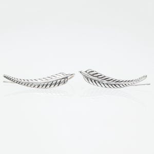 May include: A pair of silver-colored feather-shaped earrings. Each earring has a curved design with detailed feather vein patterns. The earrings are displayed on a white surface, with a slight reflection visible.