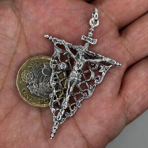 May include: A detailed silver pendant depicting the crucifixion of Jesus Christ. The pendant features a crucifix with a figure of Christ and is set within an ornate, openwork design. The pendant is shown next to a coin for size comparison.