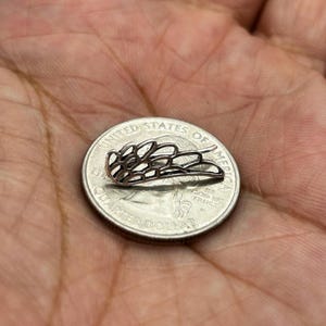 May include: A small, silver-colored metal wing-shaped earring, with a delicate openwork design, rests on a United States quarter. The earring's intricate details are visible against the coin's surface.
