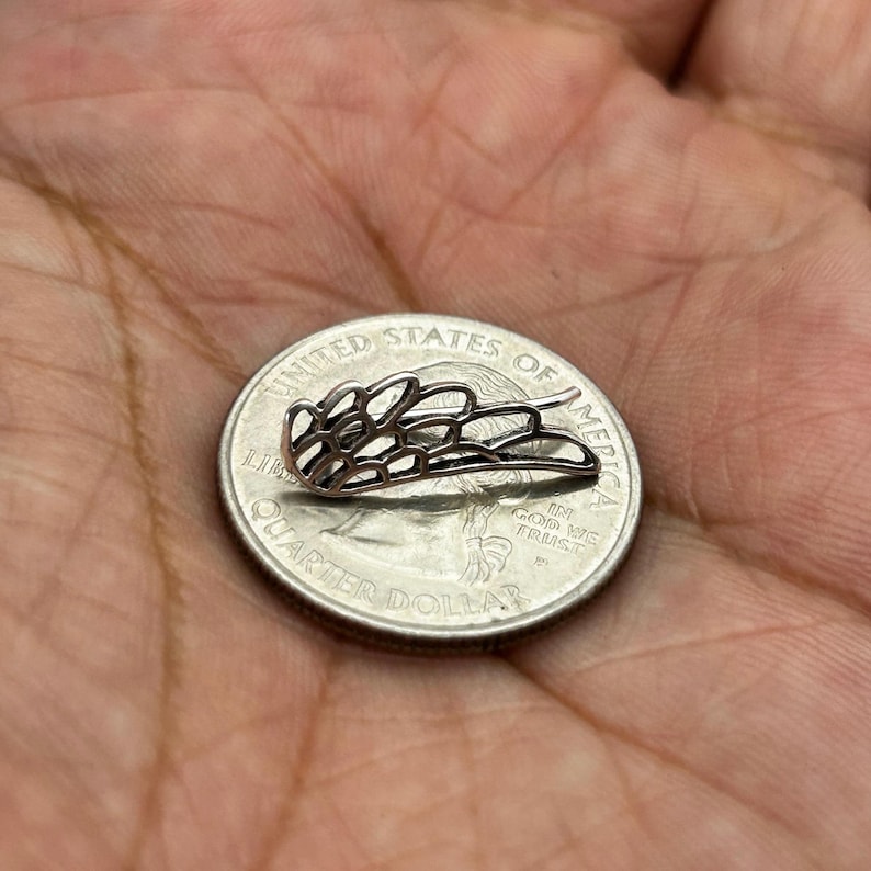 May include: A small, silver-colored metal wing-shaped earring, with a delicate openwork design, rests on a United States quarter. The earring's intricate details are visible against the coin's surface.