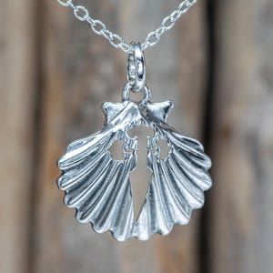 Könnte beinhalten: Eine silberne Anhänger-Halskette mit einem Jakobsmuschel-Design und einem ausgeschnittenen Kreuz. Der Anhänger hängt an einer feinen Kette. Die Muschel hat detaillierte Rippen und eine polierte Oberfläche. Der Hintergrund ist eine verschwommene Holzoberfläche.