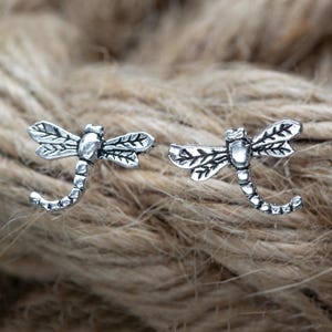 Op de afbeelding: Twee zilverkleurige oorringen in de vorm van een libelle. Elke oorring heeft gedetailleerde vleugels en een gebogen staart. De sieraden zijn geplaatst tegen een getextureerde, neutraal getinte achtergrond, die het ontwerp benadrukt.