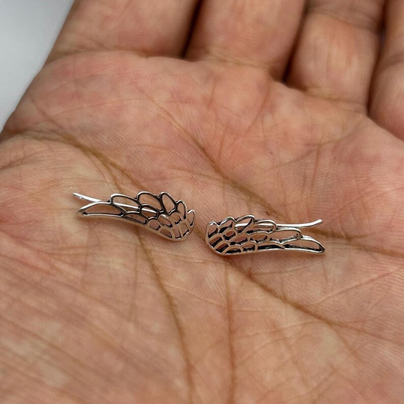 May include: A pair of silver-colored wing-shaped earrings. The earrings are designed with intricate details, resembling feathers. The wings are displayed on a hand, showcasing their size and design.