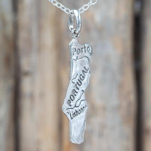 May include: A silver-toned metal pendant shaped like the map of Portugal, suspended from a delicate silver chain. The pendant features engraved text reading Porto, Portugal, and Lisbon, with embossed lines representing regional borders.