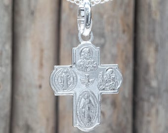 May include: A silver-colored pendant necklace featuring a cross design. The cross has religious figures and symbols, including a dove, and is suspended from a delicate chain. The background is a blurred wooden surface.
