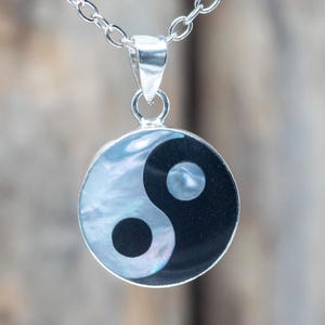 Könnte beinhalten: Eine silberne Halskette mit Anhänger, die ein rundes Yin-und-Yang-Symbol zeigt. Der Anhänger hat ein schwarzes und schillerndes weißes Perlmutt-Design. Die Halskette besteht aus kleinen, verbundenen Silberstücken.