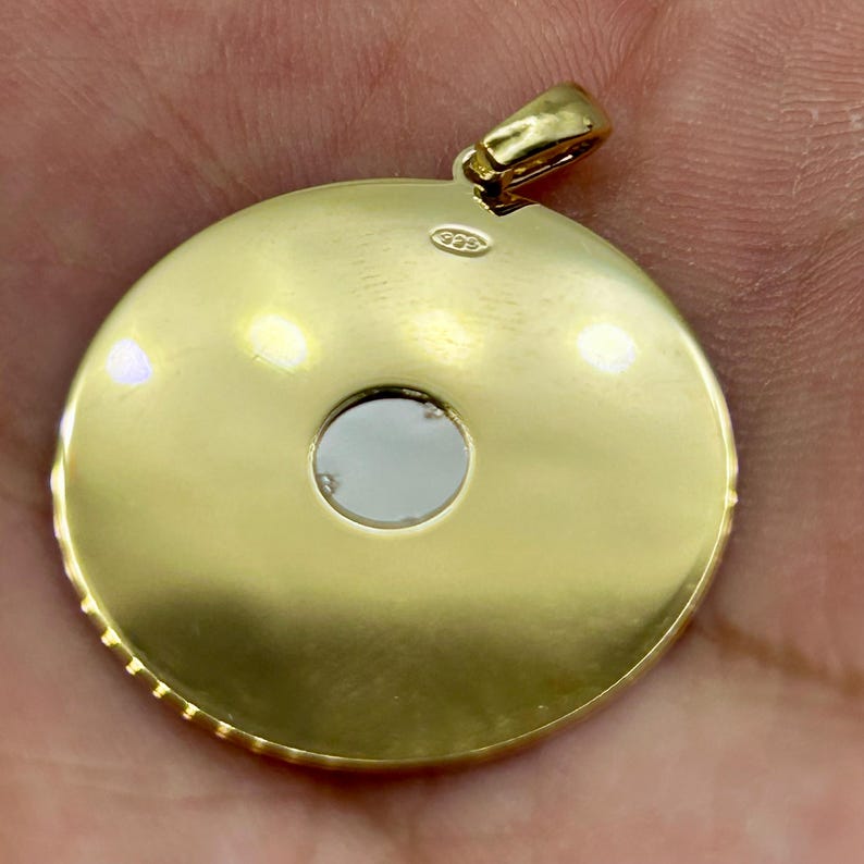 May include: A gold pendant with a circular design and a small, central opening. The pendant has a textured edge and a loop for a chain. The back of the pendant is smooth and shiny, with a small marking near the loop.