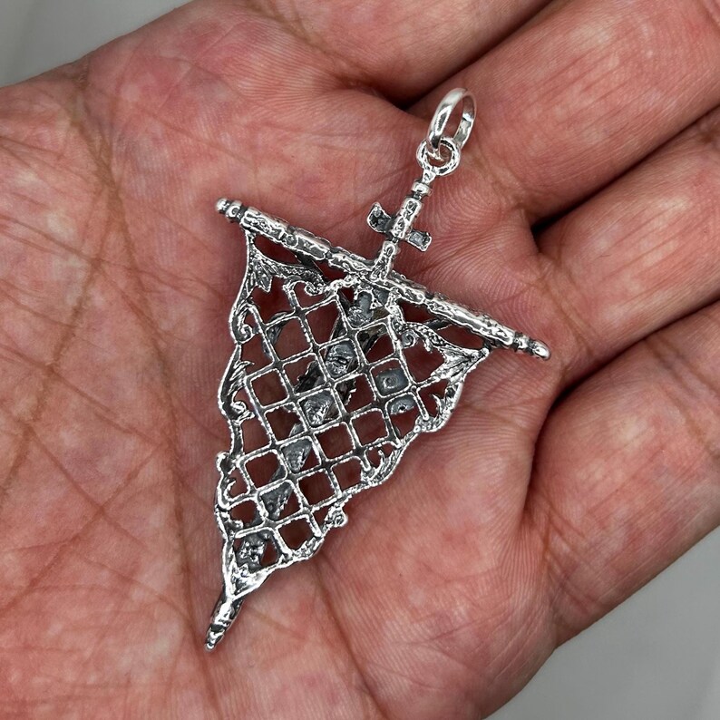 May include: A detailed silver pendant in the shape of a flag, featuring a cross and intricate openwork design. The pendant has a loop at the top for a chain. The flag design has a grid pattern with small square openings.