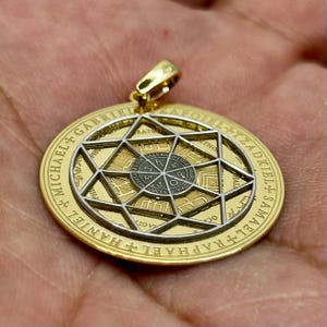 May include: Gold-tone pendant with a star-shaped design and intricate detailing. The pendant features a central circular element with symbols and text, surrounded by a larger circular frame with engraved text. A gold bail is attached at the top.