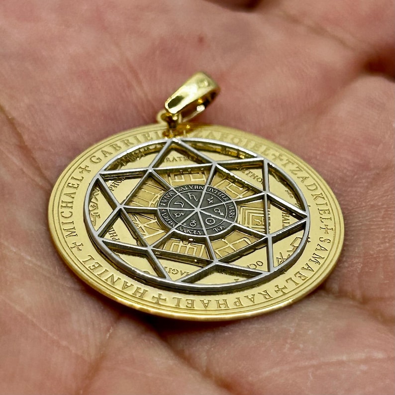 May include: Gold-tone pendant with a star-shaped design and intricate detailing. The pendant features a central circular element with symbols and text, surrounded by a larger circular frame with engraved text. A gold bail is attached at the top.