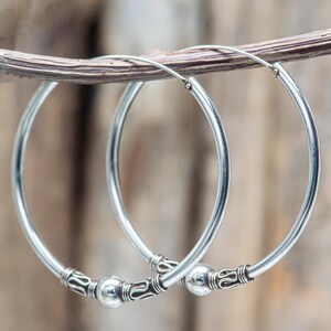Peut inclure: Une paire de boucles d'oreilles créoles argentées. Les boucles d'oreilles sont rondes avec une finition lisse et polie. Chaque boucle d'oreille comporte une perle décorative et des détails ornés au centre. Elles sont présentées sur un fond naturel flou.