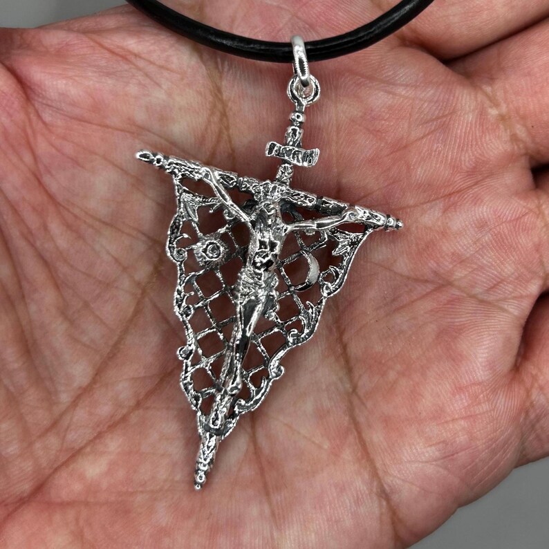 May include: A silver-colored pendant necklace featuring a detailed crucifix design. The pendant is intricately crafted with a filigree pattern and hangs from a black cord. The crucifix depicts the figure of Jesus Christ.