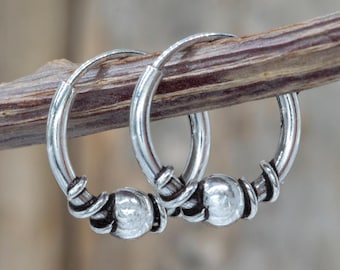 May include: A pair of silver hoop earrings. Each earring features a small, round bead and decorative wire wrapping. The earrings are displayed on a brown twig against a blurred background.