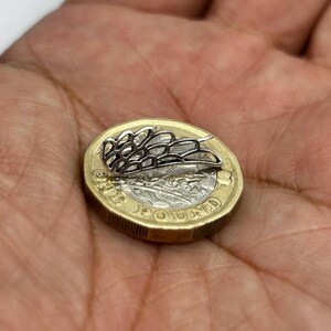 May include: A small, silver-colored, openwork wing-shaped jewelry piece rests on a gold-colored coin. The coin is a British pound, with text and details visible. The jewelry piece has a delicate, geometric design.