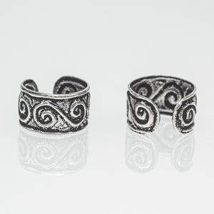 Può includere: Due anelli regolabili color argento con un design aperto. Ogni anello presenta un motivo a spirale ripetuto attorno alla fascia. Gli anelli sono esposti su una superficie riflettente, evidenziando i loro dettagli e il design.