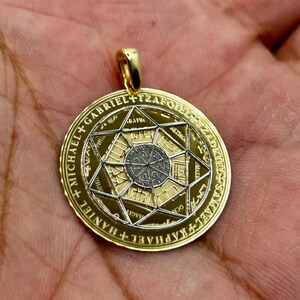 May include: Gold-tone pendant with a detailed design. The round pendant features a central geometric pattern surrounded by text and a star-shaped design. The pendant has a gold bail for attaching to a chain.