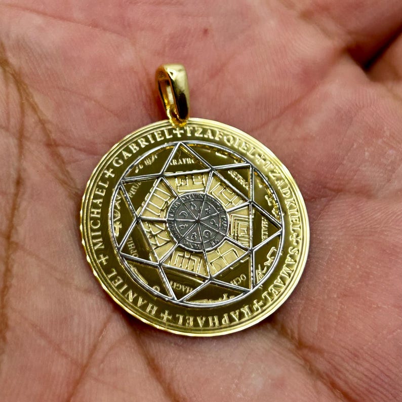 May include: Gold-tone pendant with a detailed design. The round pendant features a central geometric pattern surrounded by text and a star-shaped design. The pendant has a gold bail for attaching to a chain.