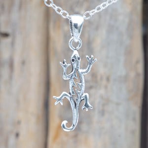 Pode incluir: Um pingente de metal prateado em forma de lagarto com um design recortado ao longo do corpo, suspenso por uma corrente de elos prateada a condizer. A figura do lagarto apresenta membros detalhados e uma cauda curva, pendurada verticalmente.