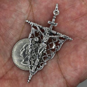 May include: A silver pendant featuring a detailed depiction of a crucified figure, with intricate filigree and a decorative banner. The pendant includes a small hammer and a loop for a chain. A quarter coin is next to the pendant for size comparison.
