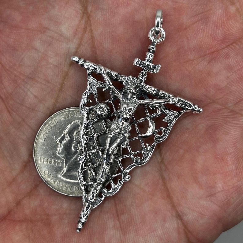 May include: A silver pendant featuring a detailed depiction of a crucified figure, with intricate filigree and a decorative banner. The pendant includes a small hammer and a loop for a chain. A quarter coin is next to the pendant for size comparison.