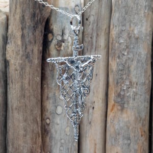 May include: A silver-toned pendant necklace featuring a detailed depiction of the crucifixion. The pendant has a banner shape with a lattice design, and a figure of Jesus Christ is visible. The necklace hangs on a delicate silver chain.