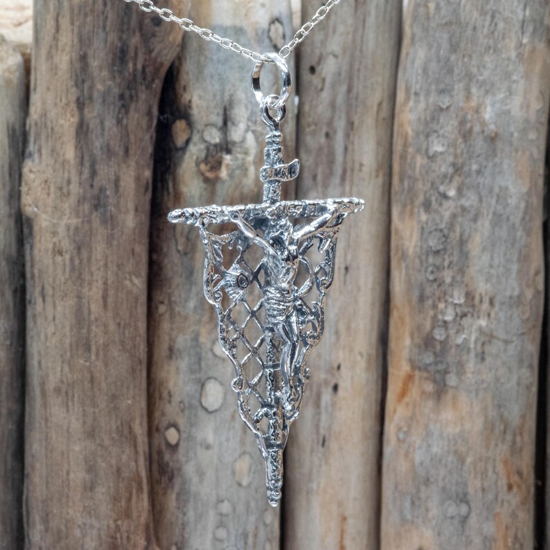 May include: A silver-toned pendant necklace featuring a detailed depiction of the crucifixion. The pendant has a banner shape with a lattice design, and a figure of Jesus Christ is visible. The necklace hangs on a delicate silver chain.