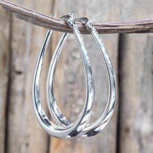 Pode incluir: Um par de brincos de argola prateados em forma de gota. Os brincos são lisos e brilhantes, com formato de gota. Estão pendurados em um galho de madeira contra um fundo de madeira desfocado. Uma joia clássica.